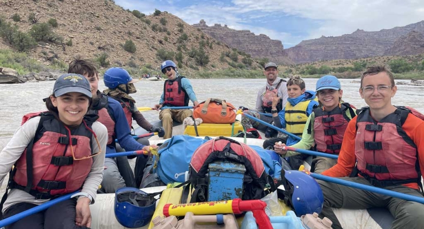 A group of people sit in a raft wearing life jackets and smile for the photo. Each person holds an oar, and there are tall canyon walls in the background. 